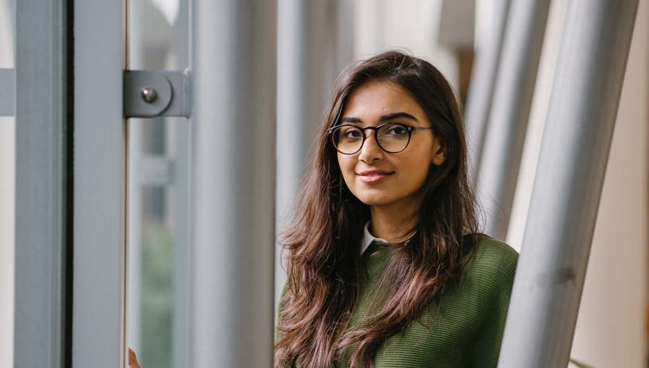 Portrait of a beautiful, young, elegant and attractive Indian Asian woman student in a preppy green sweater and spectacles. She is relaxing in a glass building during the day.