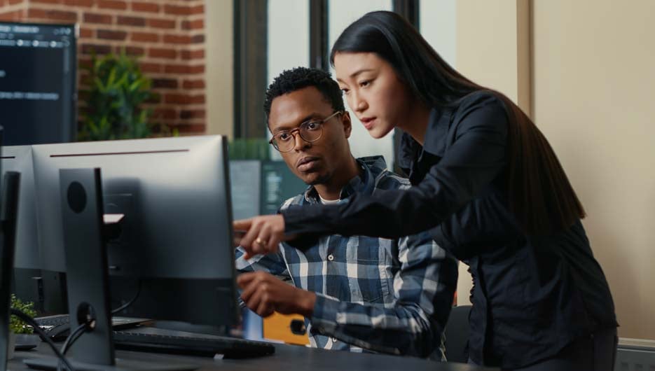 Two software developers coming at desk and sitting down holding laptop with coding interface