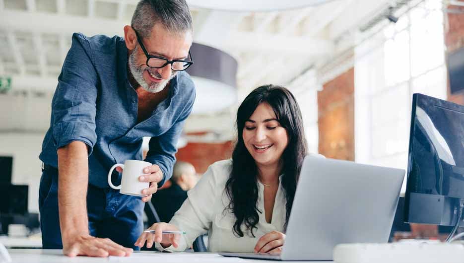 Two happy colleagues collaborating on a new project in the office. Mature businessman teaming up with his female colleague in a modern office. Two colleagues smiling while working together.